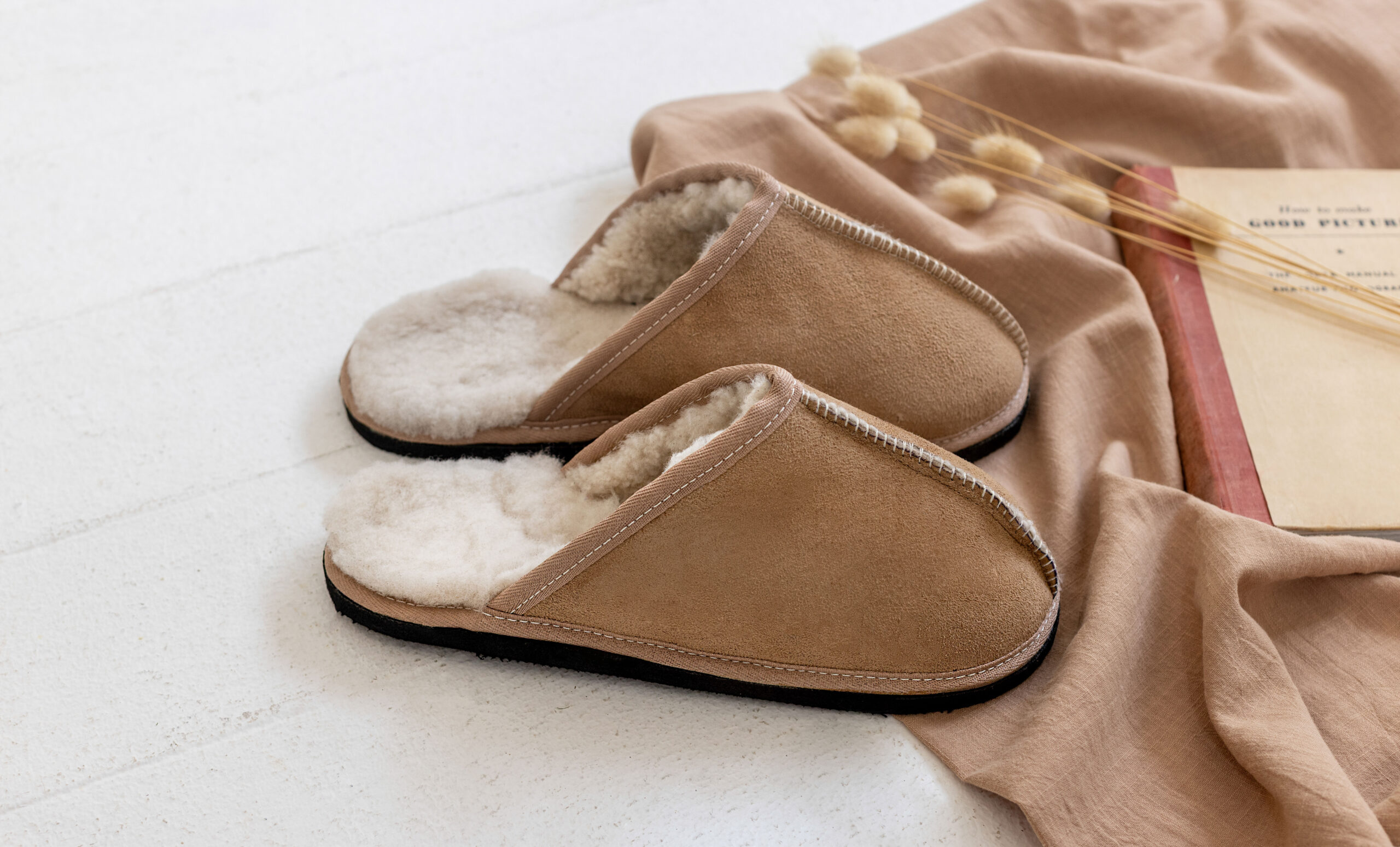 Pair of tan adult sheepskin slippers placed on a soft fabric surface beside a closed book.