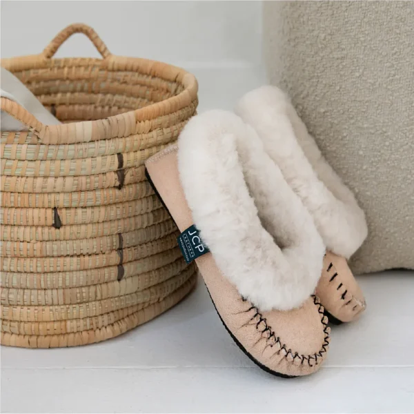 Cape Classic sheepskin slippers with visible hand-stitched detail and plush wool cuff, resting against a woven basket.