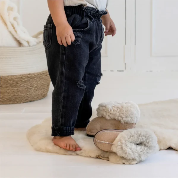 Toddler standing barefoot next to a pair of Karoo Classic junior sheepskin slippers on a soft baby rug.