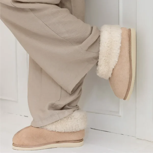 Person wearing natural sheepskin slippers with a relaxed neutral outfit, leaning against a white wall.