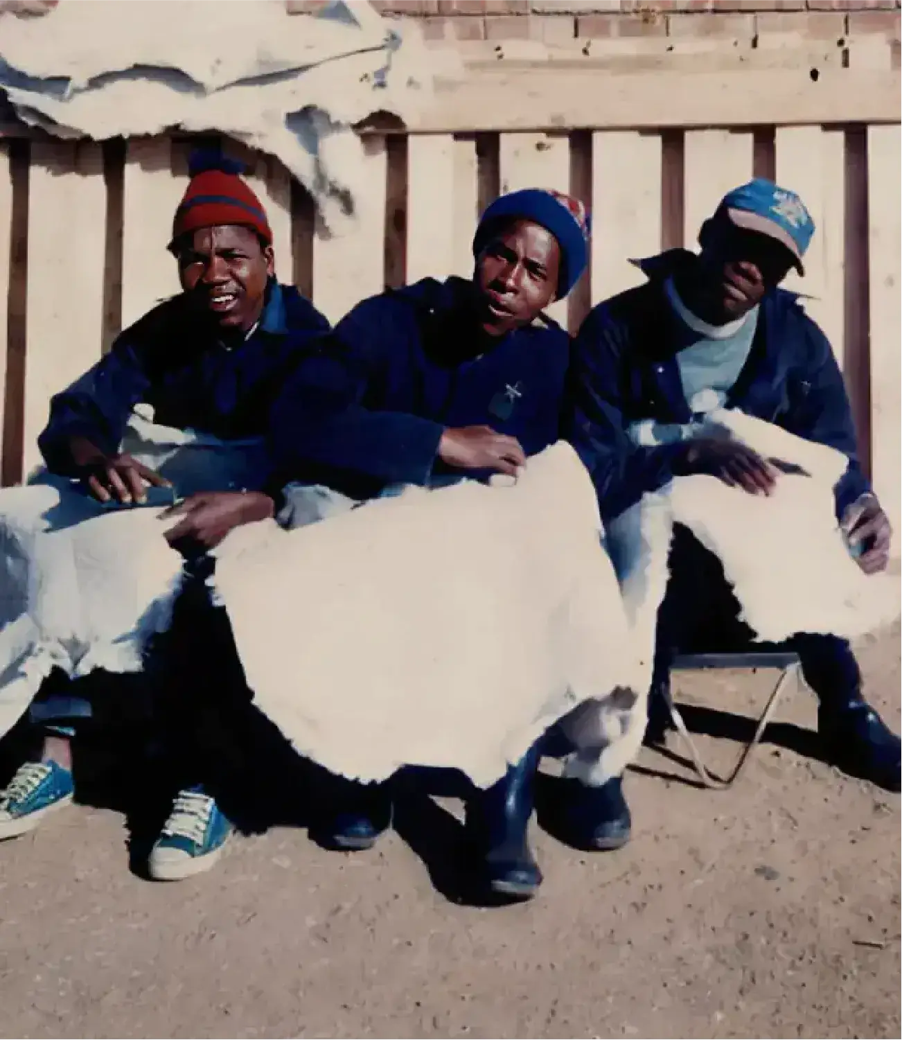 Three workers seated outdoors, each holding raw sheepskin hides during early processing stages.